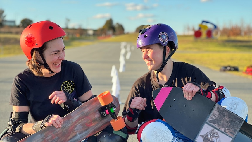 A woman wearing a red helmet holding a skateboard laughs next to another woman wearing a purple helmet