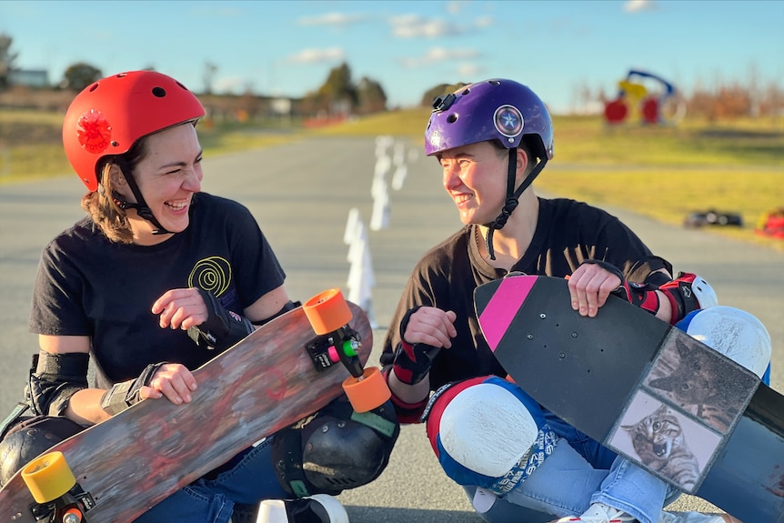 A woman wearing a red helmet holding a skateboard laughs next to another woman wearing a purple helmet