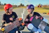 A woman wearing a red helmet holding a skateboard laughs next to another woman wearing a purple helmet