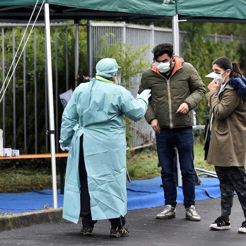 Two people wearing masks are greeted by a health worker wearing a full protective suit at a checkpoint in Italy.