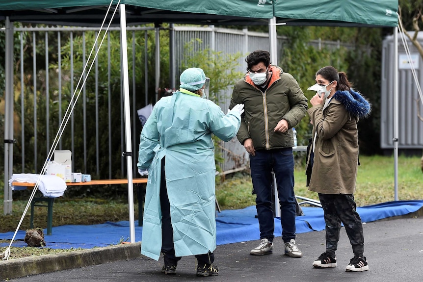Two people wearing masks are greeted by a health worker wearing a full protective suit at a checkpoint in Italy.