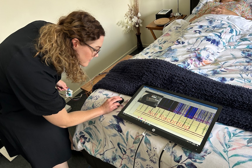 A woman with glasses and curly long hair looks at an electronic board while a female patient lies in a hospital bed