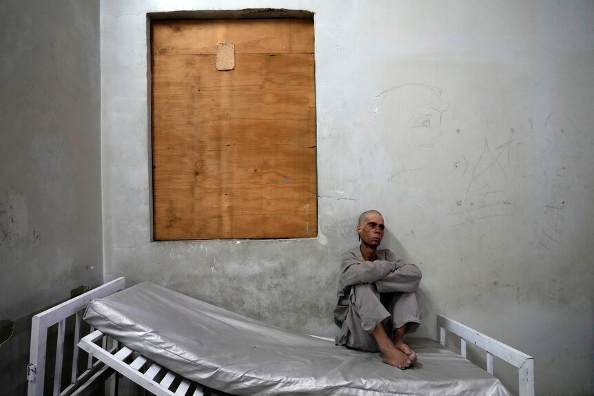 A bald, thin man in plain grey clothing sits against the wall on a bare mattress in a bare concrete room.