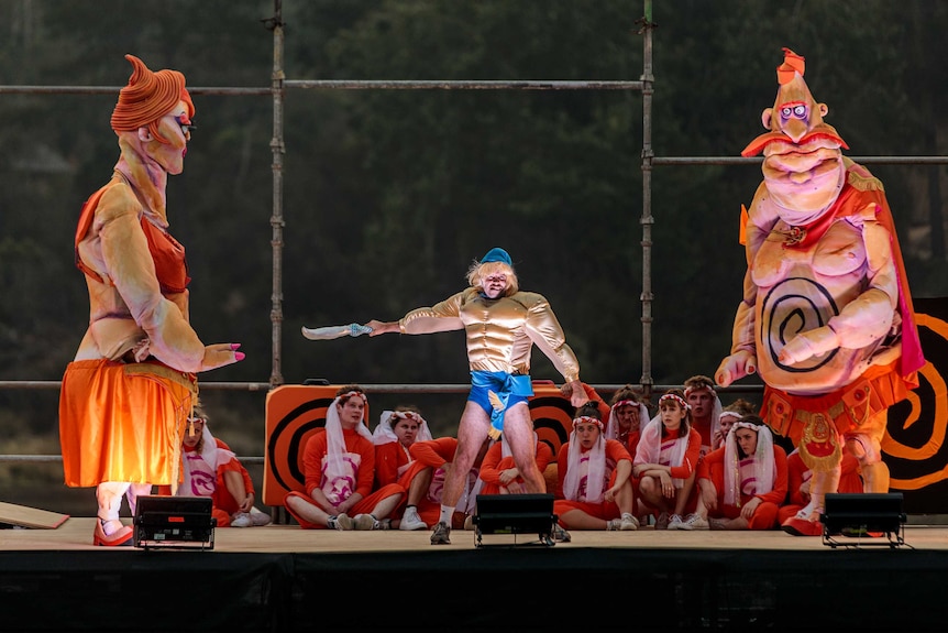 A stage with a man with comical fake abs surrounded by two enormous fantastical puppets of a man and a woman, kids in the b/g