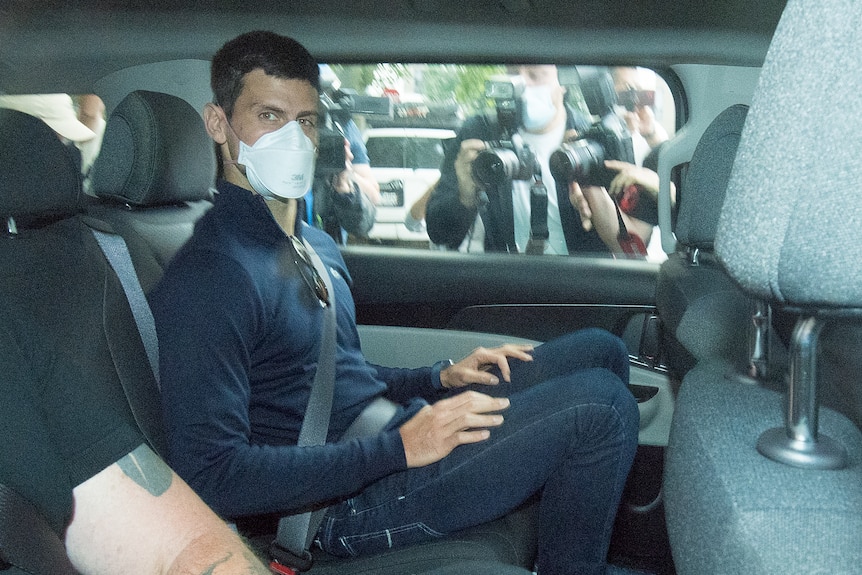 Novak Djokovic is seen sitting in the back seat of a car wearing streetwear and a face mask