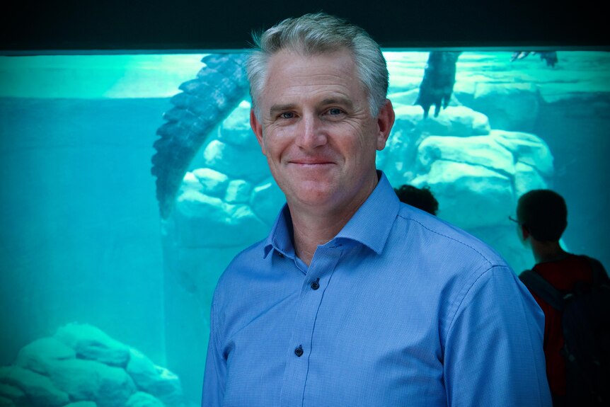 A man in a blue shirt smiles at the camera, behind him is a crocodile in a tank.