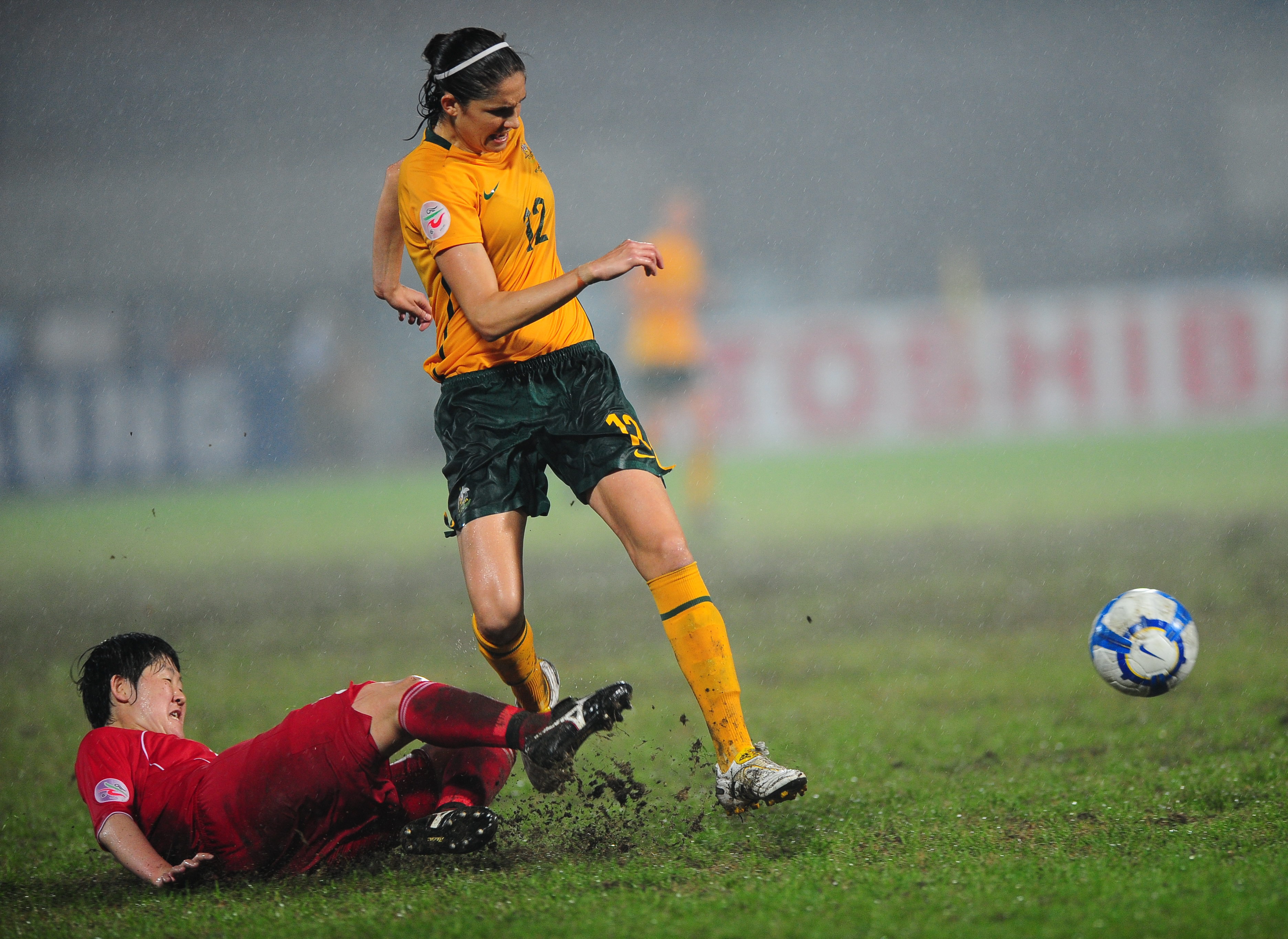 Um jogador de futebol com uniforme vermelho desliza pelo chão com a perna voltada para a bola enquanto outro jogador corre em direção a ela.