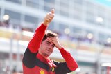 A Formula 1 driver stands at the side of the track with his hand on his forehead giving a thumbs up signal to the crowd.