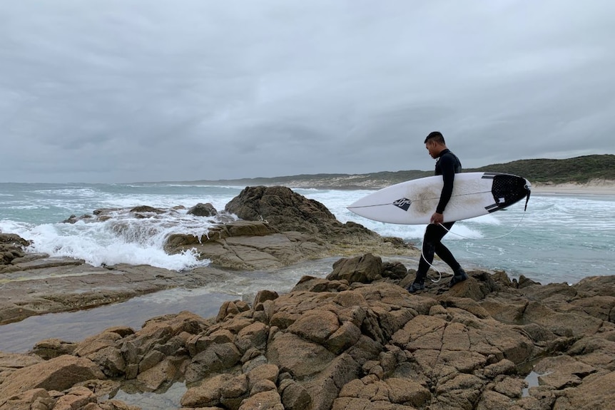 Sukma Bowling holds a surfboard and walks over rocks.