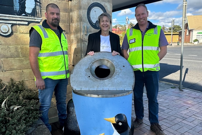 Two men and a woman stand side by side in front of a penguin-themed bin