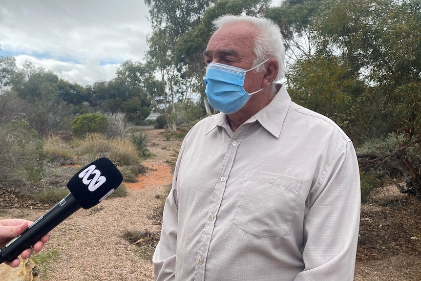 A man wearing a light coloured shirt and face mask with white hair speaks into an ABC branded microphone