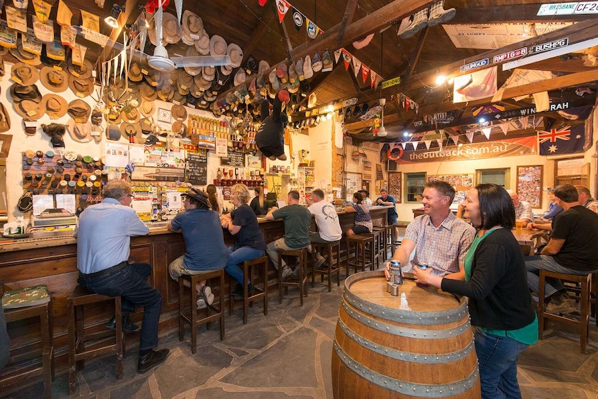 The inside of a country pub, its walls dripping with Australiana. Patrons drink at the bar.