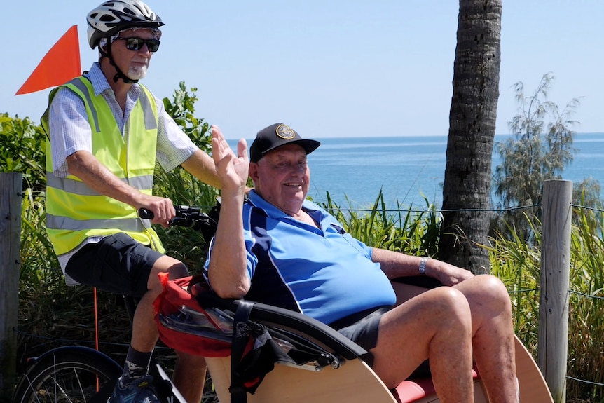 Keith sitting on the front of the trishaw waving, volunteer riding, ocean behind them.
