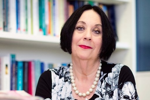 Black-haired woman sits at desk behind pile of books, holding pen and looking at camera.
