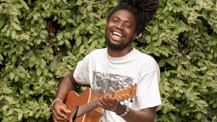 Muso looking happy playing guitar beside a hedge smiling at camera.