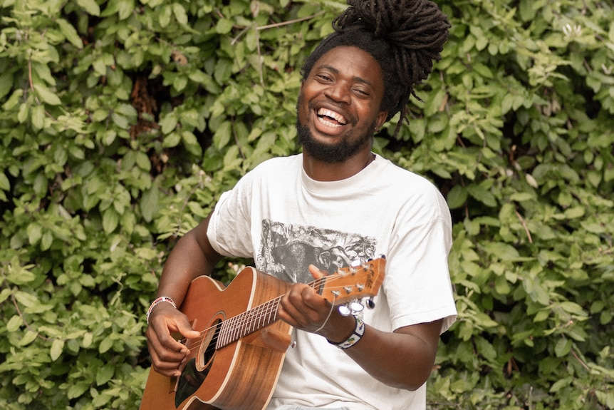 Muso looking happy playing guitar beside a hedge smiling at camera.