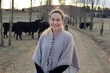 A smiling woman stands in the middle of a road in a grey shawl. A small number of cattle stand behind her.