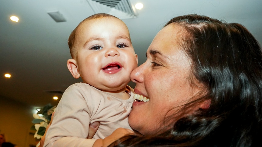 A smiling woman holds up a baby, also smiling in a tightly photographed shot taken inside.