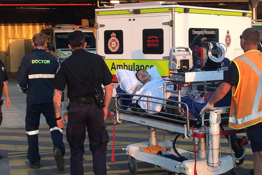 Police and paramedics walking a man on a hospital gurney behind an ambulance