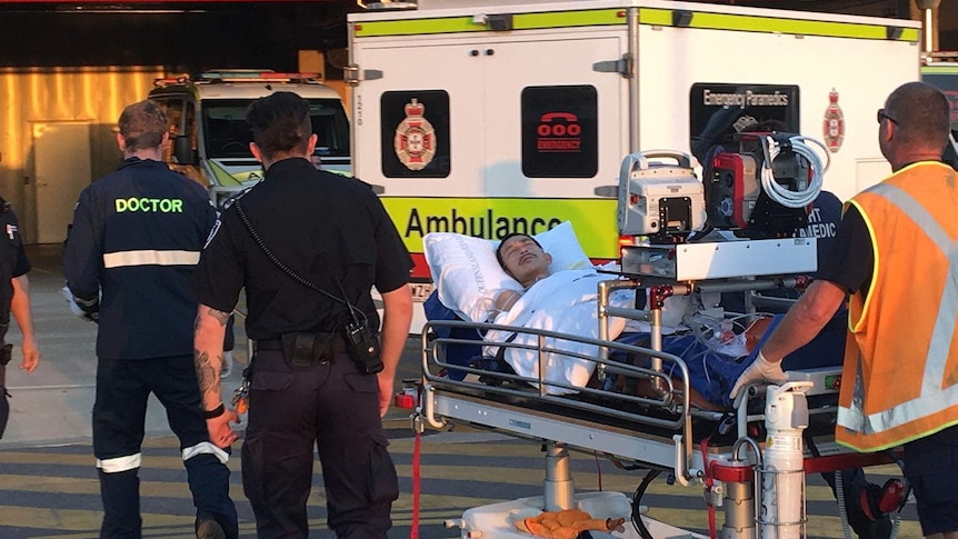 Police and paramedics walking a man on a hospital gurney behind an ambulance