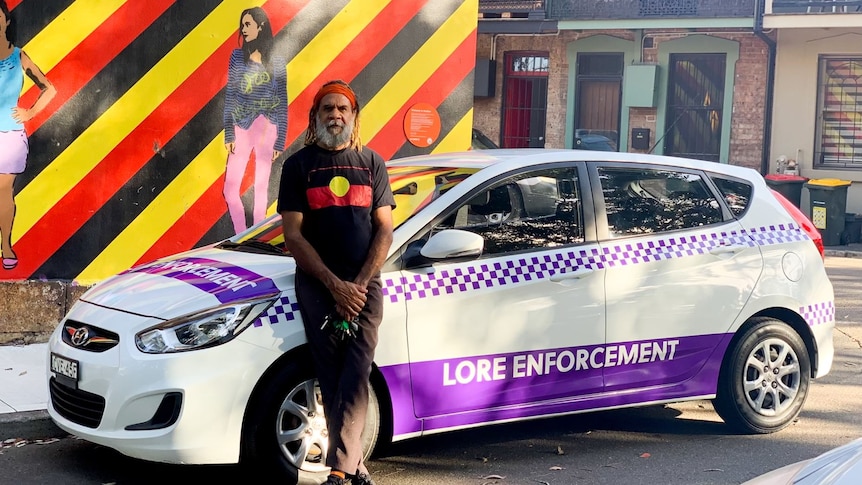 An older indigenous man wearing a shirt with an Aboriginal flag on it, standing in front of a car labelled 'Lore Enforcement'.
