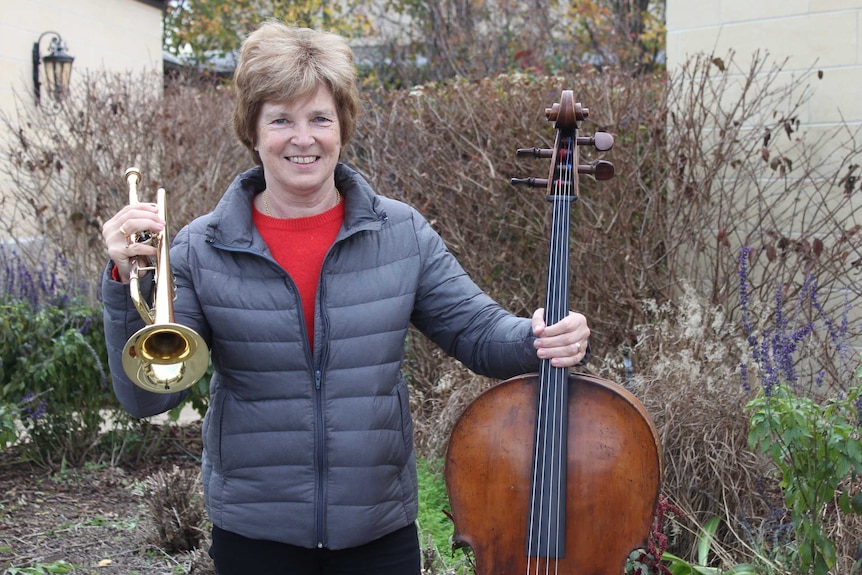 woman in a puffer jacket standing outside of a house garden holding a trumpet in right hand and cello in left hand