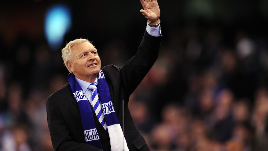A man in a dark suit wearing a football club's blue and white scarf waves at a crowd. 