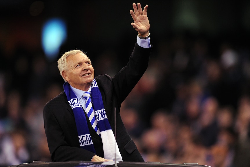 A man in a dark suit wearing a football club's blue and white scarf waves at a crowd. 