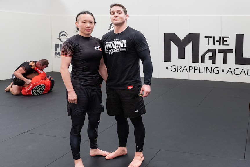 Nick and Jackie Hughes stand on a mat in a gym while two couples grapple on the floor behind them.