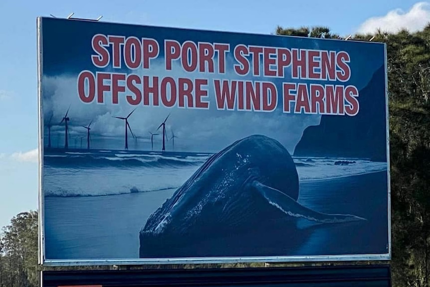 A billboard sign displays an illustration of a dead whale on a beach, with a wind farm in the background.