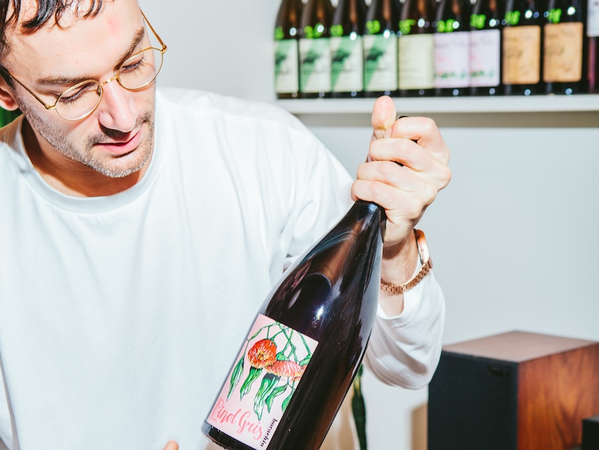 Young man with glasses holds bottle of red wine with more bottles in background 