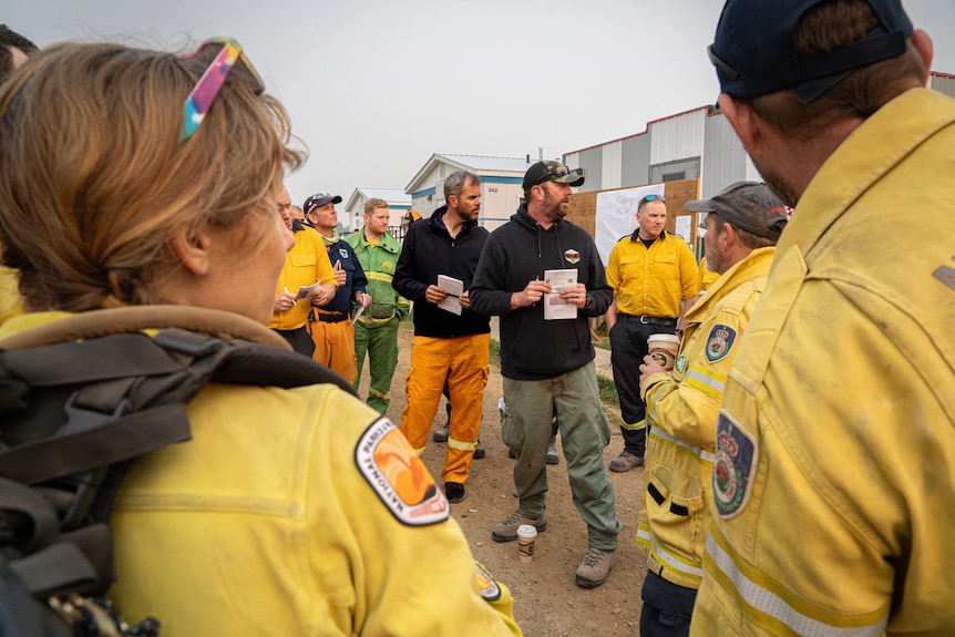 A group of firefighters stand in a circle near a billboard having a meeting under a smoky sky.
