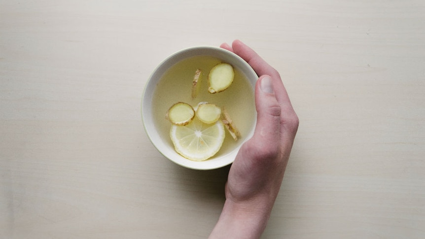 A hand cradles a mug with slices of lemon and ginger in hot water.