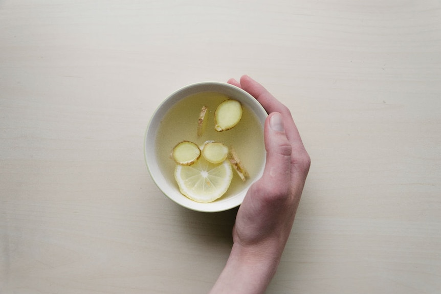 A hand cradles a mug with slices of lemon and ginger in hot water.