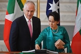 Aung San Suu Kyi writes at a lectern standing next to Malcolm Turnbull. Behind them are flags of Myanmar and Australia