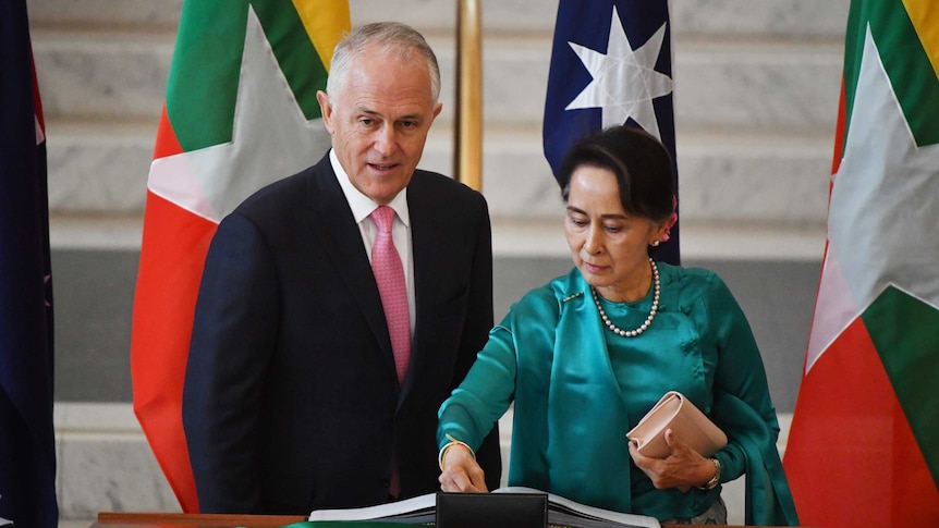 Aung San Suu Kyi writes at a lectern standing next to Malcolm Turnbull. Behind them are flags of Myanmar and Australia