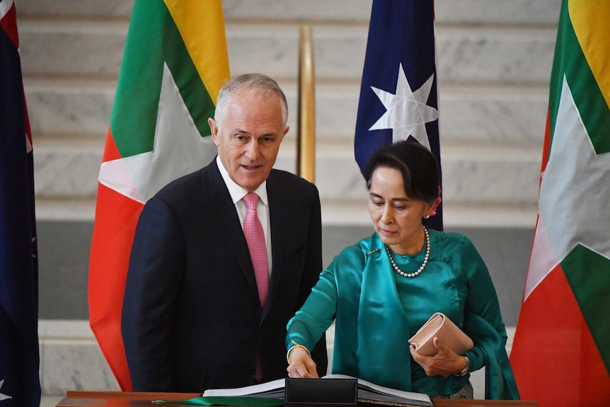 Aung San Suu Kyi writes at a lectern standing next to Malcolm Turnbull. Behind them are flags of Myanmar and Australia