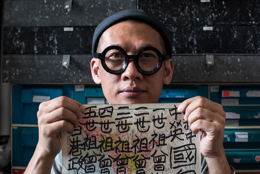 A man with thick rimmed dark glasses and a black beanie holding up a map covered in black calligraphy.