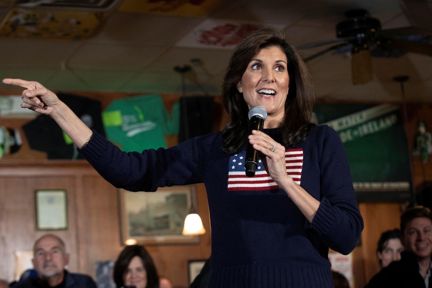 Nikki Haley, wearing a top displaying an American flag, speaks into a microphone in front of a wooded wall and a small crowd.