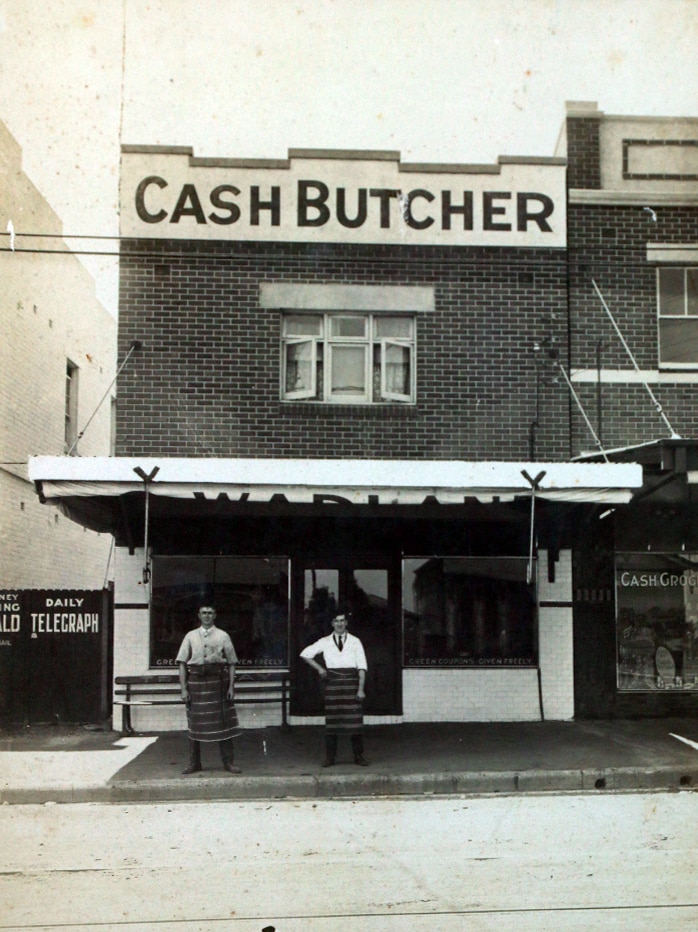 Old photograph of two men wearing aprons standing in front of a building with 'Cash Butcher' sign on the top.