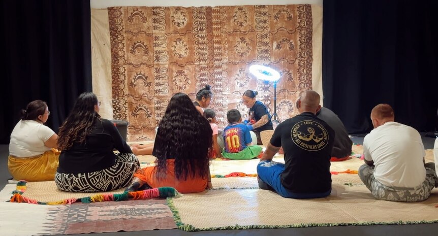 Men and women sit on fala, backs to camera as they watch tufuga in tatau session with 'au.