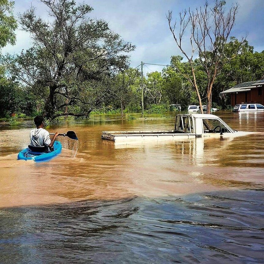 A person in a canoe paddles through floodwater that is up to the roof of a car