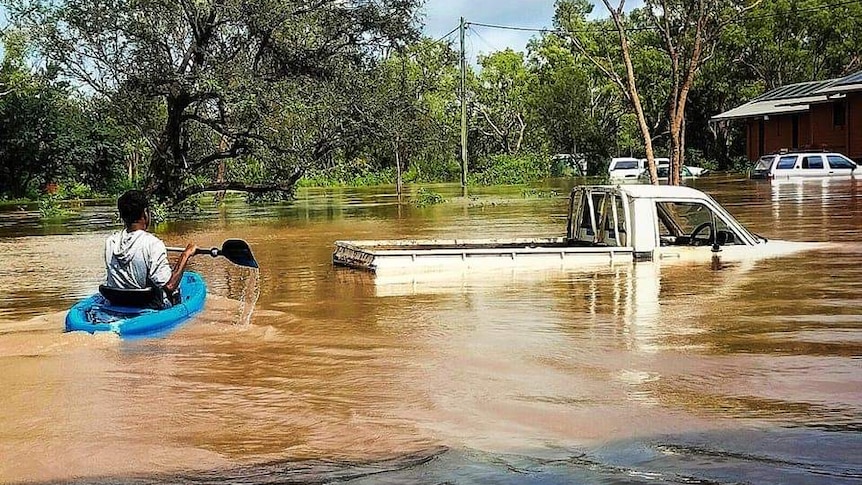 A person in a canoe paddles through floodwater that is up to the roof of a car