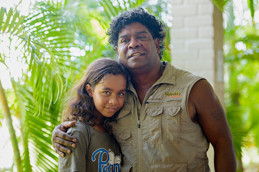 A young girl smiles at the camera, her dad's arms are around her as he looks slightly off to the distance.