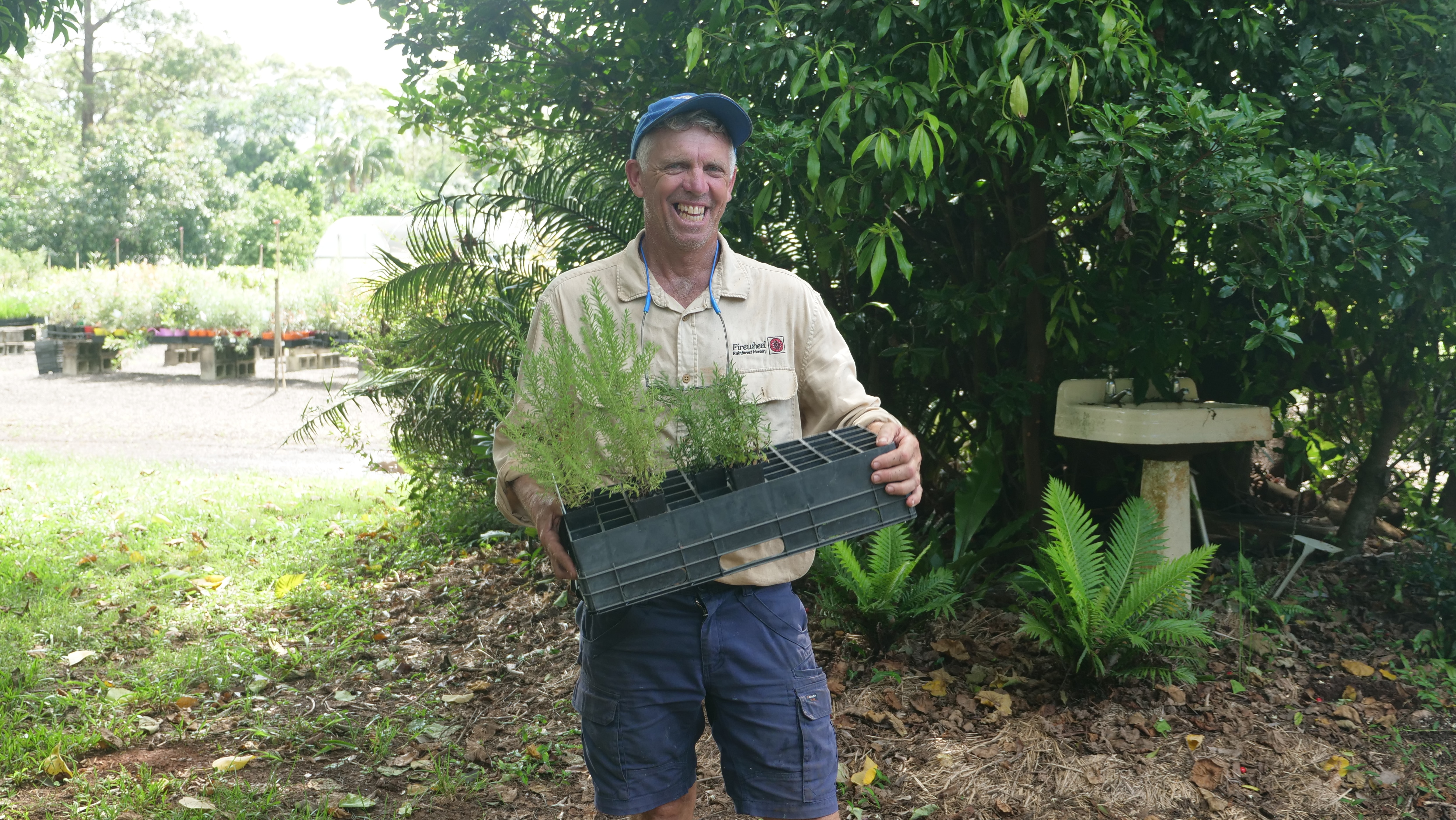 A man in outdoor workwear stands in front of a nursery, holding a tray of seedlings and laughing.