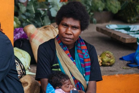 A ni-Vanuatu woman holds a baby in a marketplace surrounded by mats and vegetables