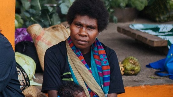 A ni-Vanuatu woman holds a baby in a marketplace surrounded by mats and vegetables