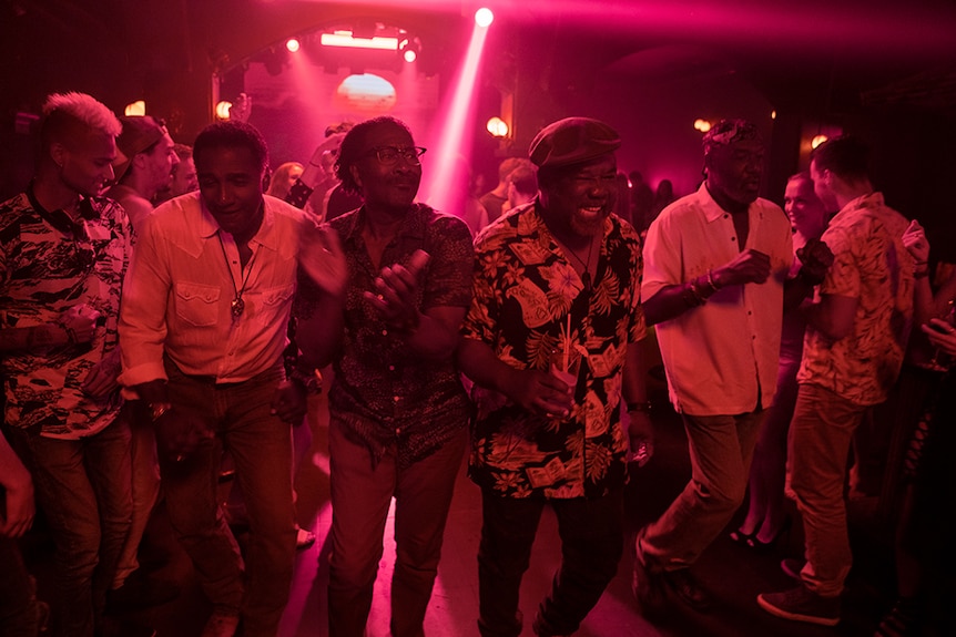 Four middle-aged men wear plain and patterned button up shirts and walk in a line through dark and busy pink-hued nightclub.