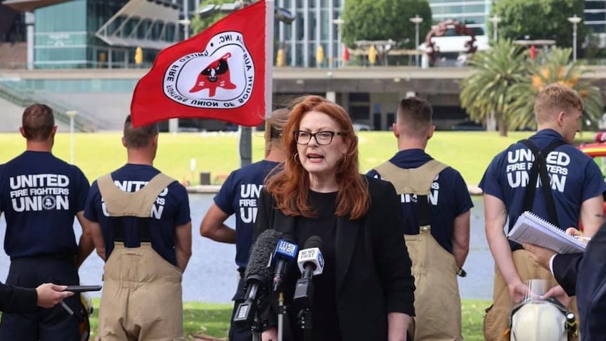 A woman stands in front of people with red flags wearing black.