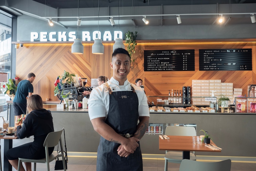 A man of Filipino heritage in white shirt and black apron standing in front of food counter in cafe.